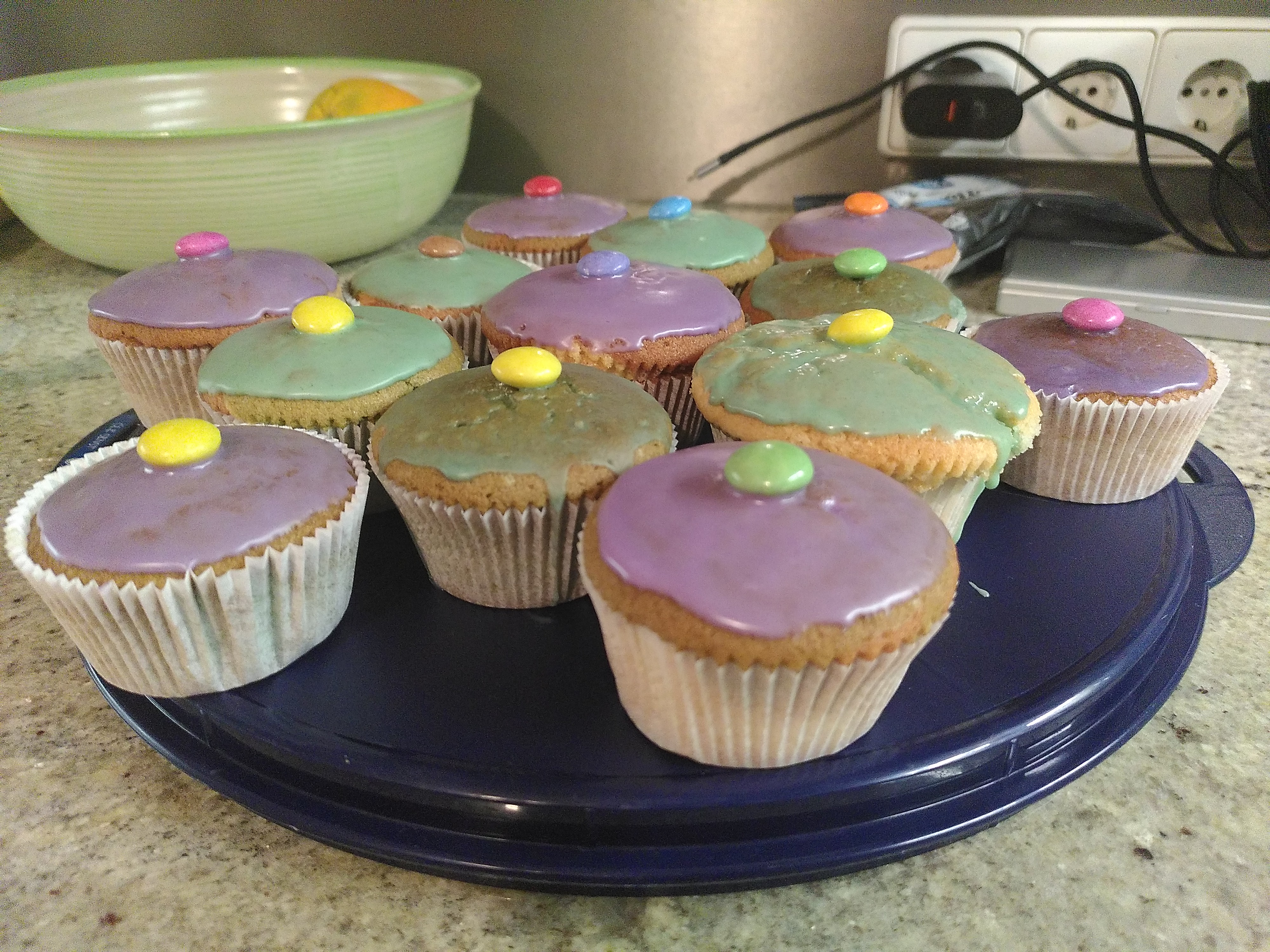 12 Muffins mit lila und grünem Zuckerguss uns jeweils einem bunten Smartie in der Mitte, stehen auf einer blauen Kuchenform aus Plastik.