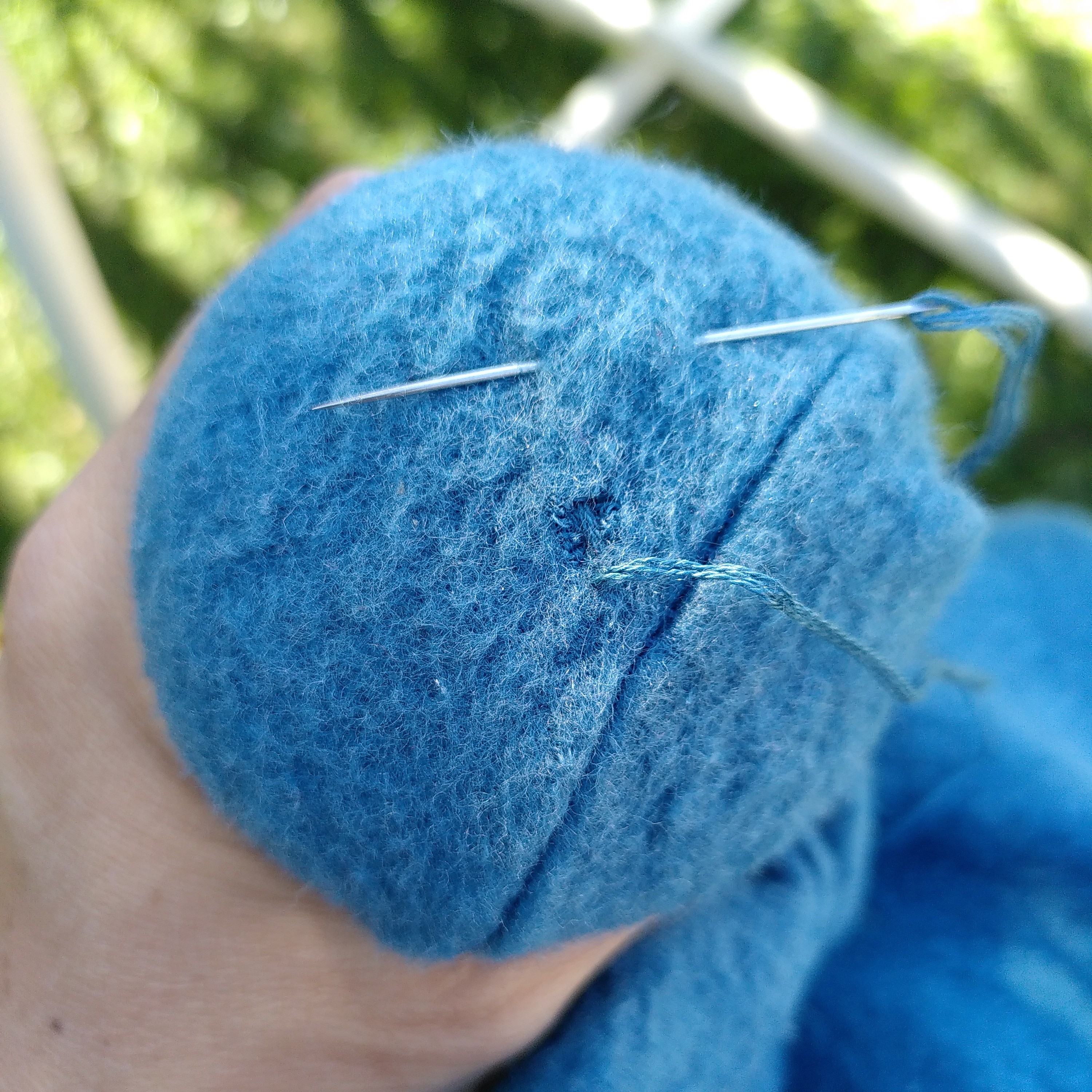Verschlossenes Loch in einem hellblauen Wollfleeceschlafsack. Man sieht, dass ein Stopfpilz unter dem Soff liegt und eine Hand diesen festhält. Oben im Stoff steckt eine Nadel, durch die das genutzte hellblaue Stopfgarn hindurchgefädelt ist.