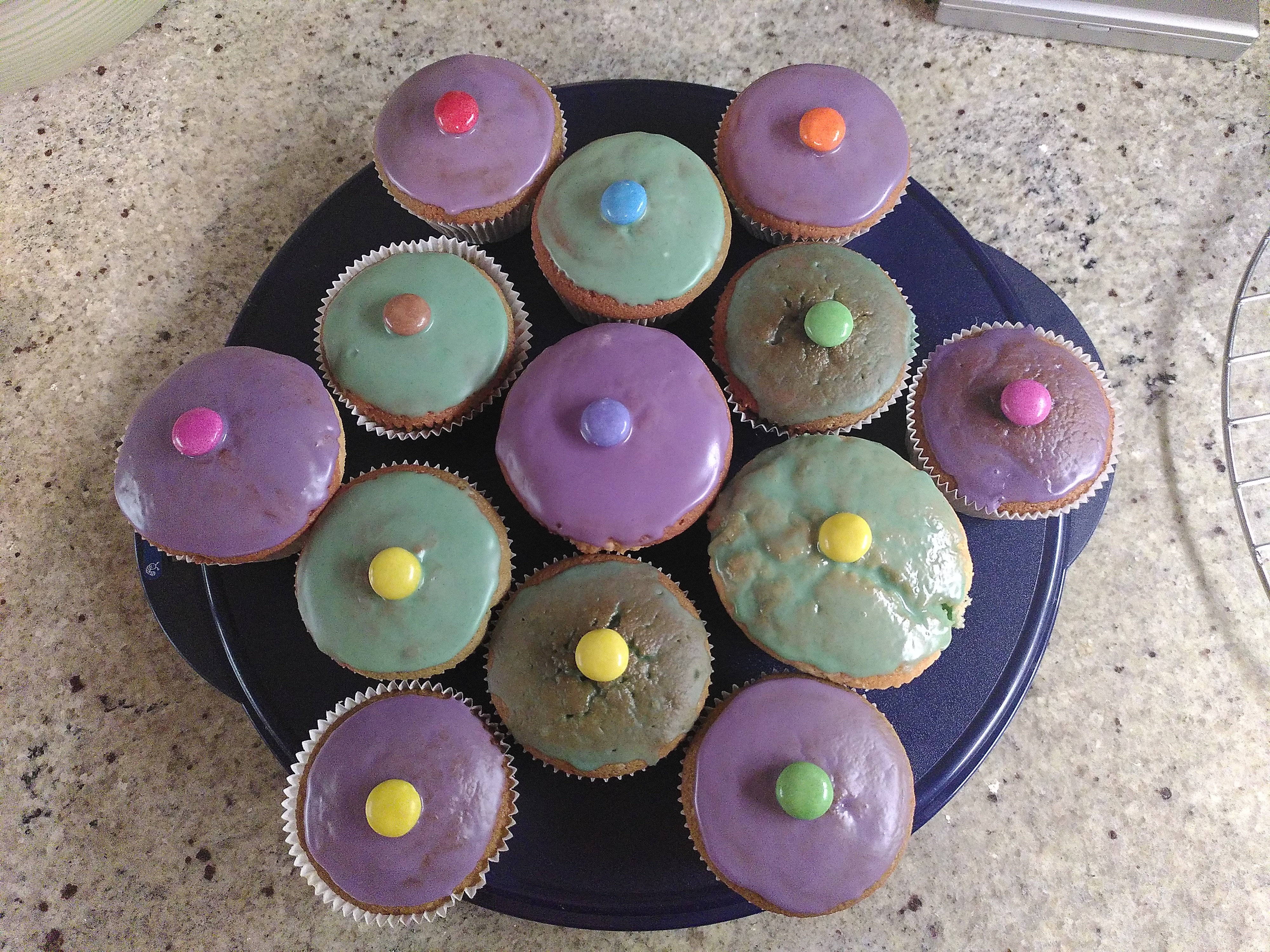 Von oben fotografiert: 12 Muffins mit lila und grünem Zuckerguss uns jeweils einem bunten Smartie in der Mitte, stehen auf einer blauen Kuchenform aus Plastik.